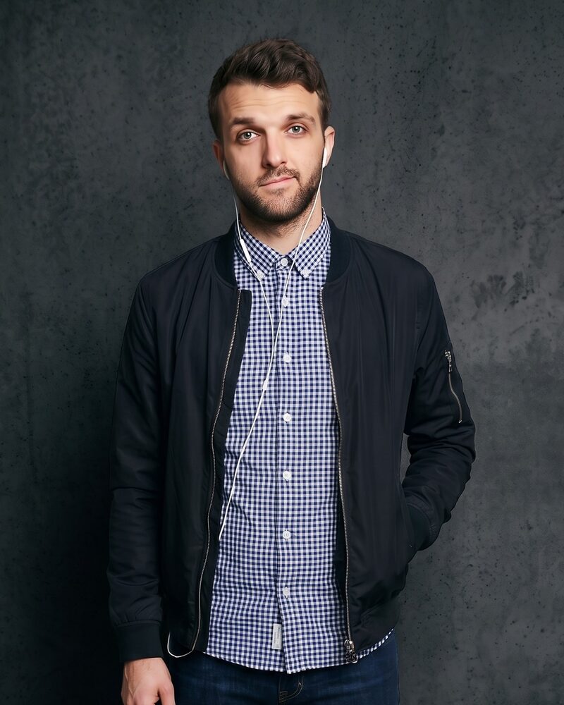 Foddy, founder of DGTL.ONE, wearing a dark bomber jacket and checked shirt in front of a dark grey concrete wall